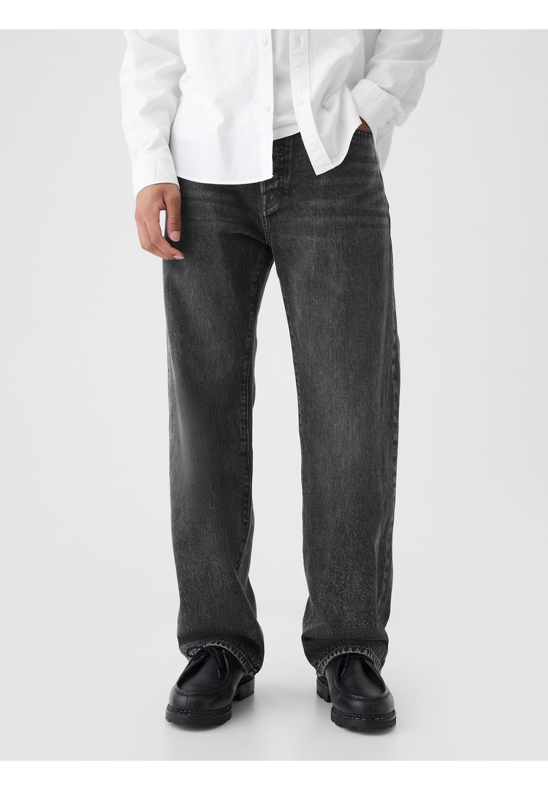 Jean en denim noir à coupe ample, présentant un léger délavage et un design classique à cinq poches. Porté avec des chaussures noires et une chemise blanche.