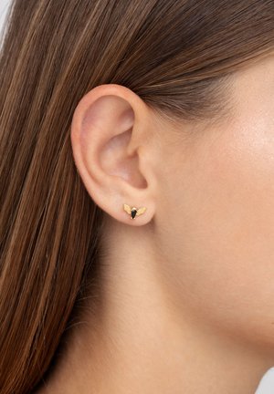 Orecchini a bottone tono oro a forma di ape, con accenti di pietre nere e superfici lisce, indossati all'orecchio di un modello.