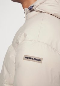 Beige puffer jacket with a hood, featuring a smooth texture. Black "PEGADOR" label on the left shoulder. Zipper hardware detail visible.