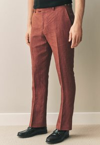 Pantalons en lin de couleur rouille, ajustés, avec une texture lisse, un devant plat et des poches latérales, associés à des mocassins noirs.