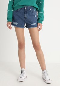 Pantalones cortos de mezclilla con un diseño desgastado, dobladillo deshilachado y lavado azul claro. Combinados con zapatillas altas blancas y una blusa de manga larga verde.