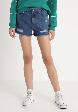 Denimshorts med et slidt design, frynset kant og lyseblå vask. Sammen med hvide højtosede sneakers og en grøn langærmet top.