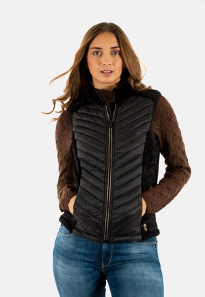 Vrouw die een zwarte doorgestikte bodywarmer draagt over een bruine gestructureerde top met lange mouwen en een blauwe spijkerbroek, staand met handen in de vestzakken.