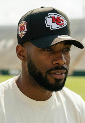 Mann mit Bart, der eine schwarze Kansas City Chiefs Mütze mit 50-jährigem Jubiläums-Patch und ein weißes Rundhalsshirt trägt, draußen bei Tageslicht.