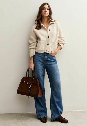 Cardigan côtelé crème avec des boutons noirs, associé à un jean bleu à jambes larges. Sac à main en daim marron avec un accent porte-clés et des chaussures marron foncé.