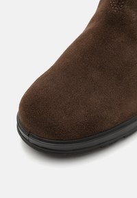 Bota de tobillo de ante marrón con punta redonda, superficie texturizada y suela de goma negra. Presenta costuras visibles y un ligero tacón.