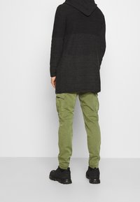 Cardigan nero lungo con cappuccio e motivo testurizzato, indossato sopra pantaloni cargo verde oliva con tasche laterali, abbinato a scarpe nere.