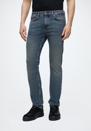 Homme portant un jean en denim bleu ajusté et des baskets noires debout devant un fond uni.