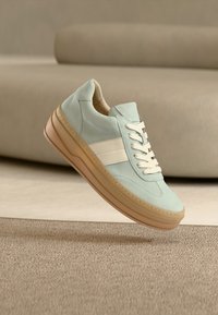 Baskets en daim vert clair avec des rayures blanches, des lacets beige, et une semelle épaisse couleur tan sur un tapis beige près d'un canapé assorti.