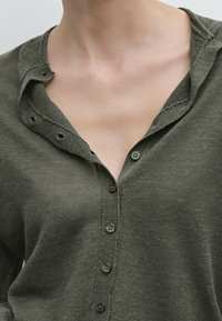 Oliven grøn skjorte med knapper lavet af tekstureret stof, der har en krave med udskæringsdetaljer og seks matchende knapper foran.