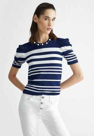 Top a righe navy e bianche a maniche corte con spalle a sbuffo e decorazioni simili a perle attorno al colletto. Abbinato a pantaloni bianchi a vita alta.
