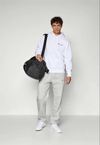 Witte hoodie met een logo, grijze sweatpants en witte sneakers. Houdt een zwarte sporttas vast. Eenvoudig ontwerp met een relaxte pasvorm.