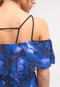 Blå blommig off-shoulder topp med svarta remmar, smockad midja och genomskinligt tyg med livliga blombladsmönster.