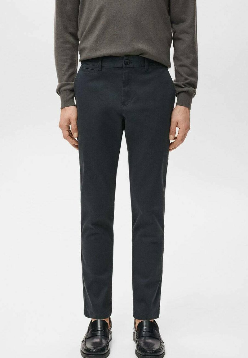 Mężczyzna ubrany w czarne, dopasowane spodnie, ciemnobrązową koszulę z długim rękawem oraz czarne skórzane mokasyny, stojący na jednolitym tle.