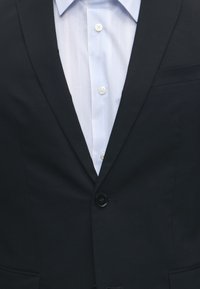 Giacca da completo nera abbottonata con un bottone sopra una camicia bianca con colletto a righe verticali e bottoni visibili.