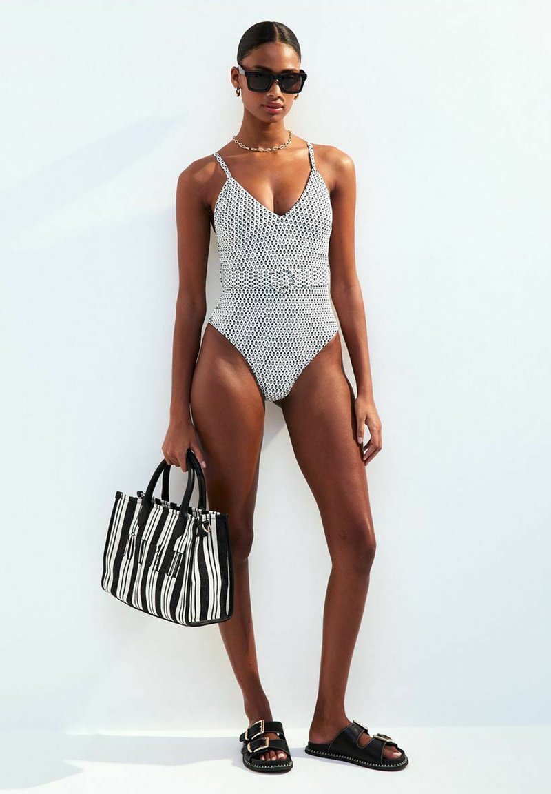 Maillot de bain une pièce à pois noir et blanc avec un décolleté en V profond, taille cintrée et fines bretelles. Accessoires comprenant un sac à main rayé noir et des sandales.