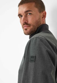 Mann mit kurzem braunem Haar und Bart, der eine dunkelgraue Jacke mit dem Brax-Logo auf dem Oberarm trägt und über seine Schulter schaut.