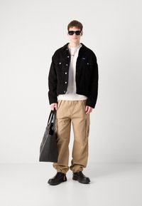Black corduroy jacket, white sweater, beige cargo pants, black glossy shoes, and a large black tote bag. Sleek sunglasses complete the look.