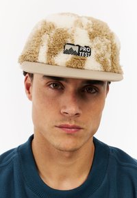 Beige and cream plush-textured cap with a flat brim, featuring a black Protest logo patch on the front. Worn by a person with short hair.