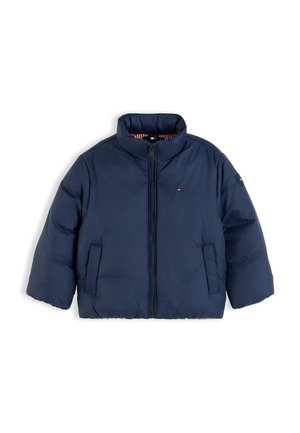 Ragazzo giovane in piedi con le mani nelle tasche che indossa una giacca imbottita blu navy, camicia bianca a righe, jeans blu larghi e sneakers bianche su sfondo bianco.