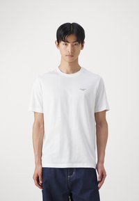 Witte katoen t-shirt met een ronde hals, korte mouwen en een klein zwart logo op de linkerkant van de borst. Model draagt donkere denim jeans.