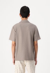 Camisa de manga curta, com gola, em castanho suave com riscas horizontais subtis. O tecido parece leve e com uma textura suave.