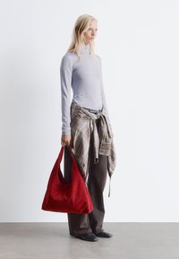 Rote Wildleder-Hobo-Tasche mit geschwungener Form; Modell trägt ein helllila enges Oberteil, einen karierter Wickelmantel und dunkle Hosen mit schwarzen Schuhen.