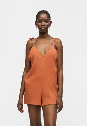 Mujer con el cabello trenzado, vistiendo un mono sin mangas color óxido con tirantes atados en los hombros y cuello halter verde oliva, sobre fondo blanco.