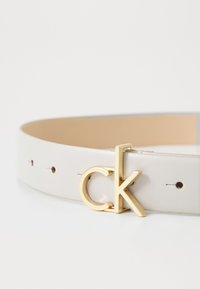 Curea din piele albă, având o cataramă cu logo-ul "ck" aurie și multiple găuri de ajustare pe curea, textură netedă și design minimalist.