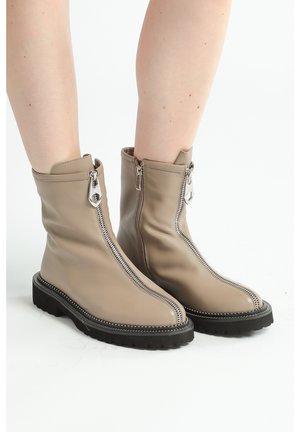 Botines de cuero beige hasta el tobillo con una cremallera plateada y suela de goma negra. Cuentan con un diseño de costura frontal único y detalles de tachuelas a lo largo de la suela.