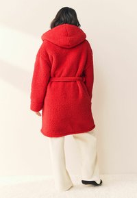 Roter, flauschiger Kapuzenmantel mit einem taillierten Gürtel, langen Ärmeln und weicher Textur. Über cremefarbigen Hosen getragen.