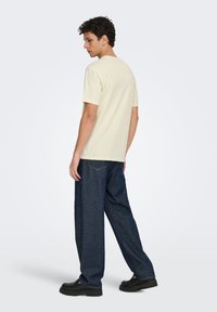 Ανοιχτό κίτρινο βαμβακερό κοντομάνικο μπλουζάκι, συνδυασμένο με φαρδιά σκούρα μπλε τζιν παντελόνια και μαύρα chunky αθλητικά παπούτσια.