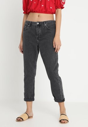 Frau trägt hochgekrempelte schwarze Jeans, eine rote bauchfreie Bluse mit Puffärmeln und beigen gewebten Slip-On-Sandalen und steht vor einem schlichten Hintergrund.