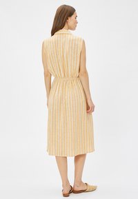 Vestido midi sin mangas de rayas amarillas y blancas con un cuello, cinturilla fruncida y tejido ligero, llevado con sandalias deslizantes beige.