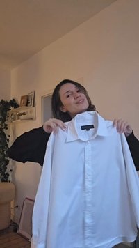 Bijela košulja s dugim rukavima, s gumbima sprijeda i velikim ovratnikom, držana ispred osobe, s opuštenim krojem i glatkom teksturom.