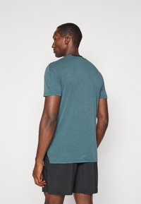 Camiseta corta de manga en color azul verdoso con un cuello redondo, hecha de una tela texturizada, que presenta un acento negro en la parte posterior. Llevada con pantalones cortos negros.