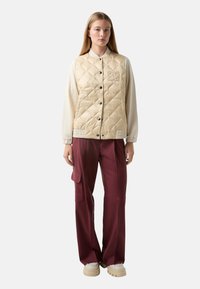 Beżowa pikowana kamizelka z zatrzaskami, z subtelnym logo, w połączeniu z burgundowymi spodniami cargo i beżowymi platformami.