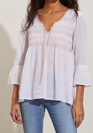 Woman wearing a white long-sleeve blouse with pleated details and a tie neckline, paired with blue jeans.