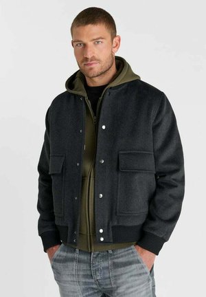 Uomo con capelli corti che indossa una giacca grigio scuro con bottoni sopra una felpa verde oliva con zip e jeans sbiaditi, mani nelle tasche.