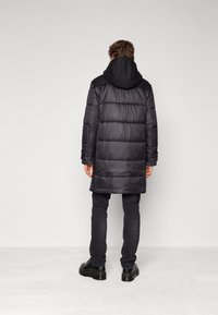 Černý puffer kabát s kapucí, s prošívaným designem a střední délkou. Nositelný přes tmavé džíny a boty, zobrazený ze zadní strany.
