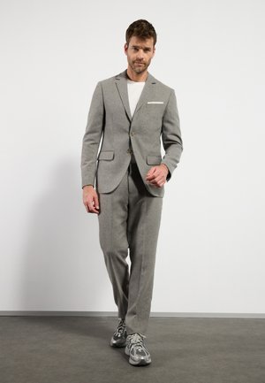 Traje gris de lana que incluye una chaqueta de un botón y pantalones ajustados, combinado con zapatillas grises. Destaca un detalle en blanco en el bolsillo de la chaqueta.
