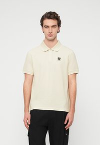 Μπεζ πόλο μπλουζάκι με γιακά, κοντά μανίκια και μικρό λογότυπο στη αριστερή πλευρά του στήθους. Ομαλή υφή, συνδ υασμένο με μαύρο παντελόνι.