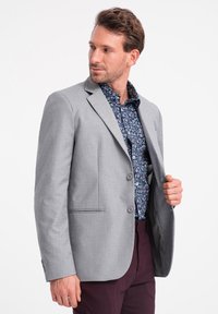 Harmaat bleiseri, jossa on sileä pinta, kaksi nappia ja yksi rintatasku, kerroksittain tummansinisen kukkapaitan ja viininpunaiset housut päällä.