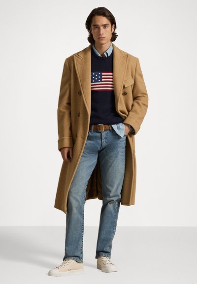 Beige ullkåpe, marineblå genser med amerikansk flaggdesign, lys blå skjorte, blå jeans og beige joggesko. Løs lagdelt antrekk.