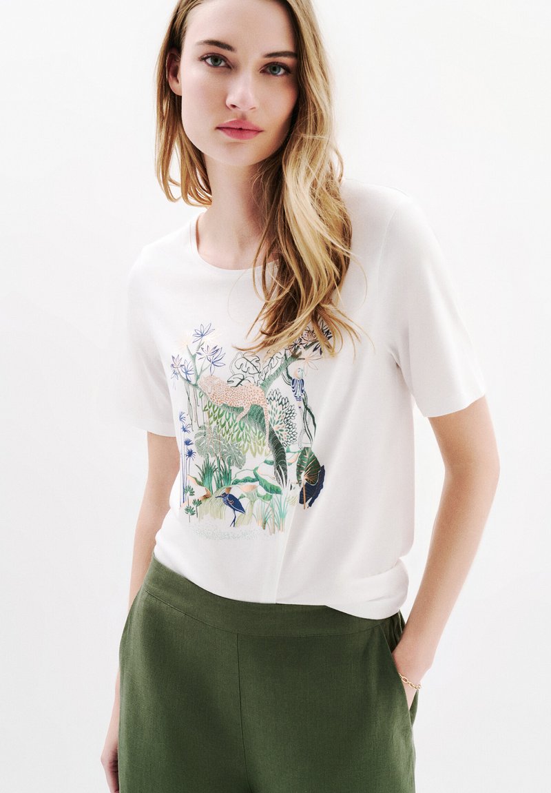 Camiseta blanca de manga corta con un diseño gráfico de temática natural en verde y azul. Combinada con pantalones verde oliva, con una textura de tela suave.