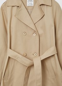 Beige, doppelt geknöpfter Mantel aus Baumwolle mit großen Knöpfen, einem Gürtel in Taillenhöhe und einem Reverskragen. Zwei seitliche Taschen sichtbar.