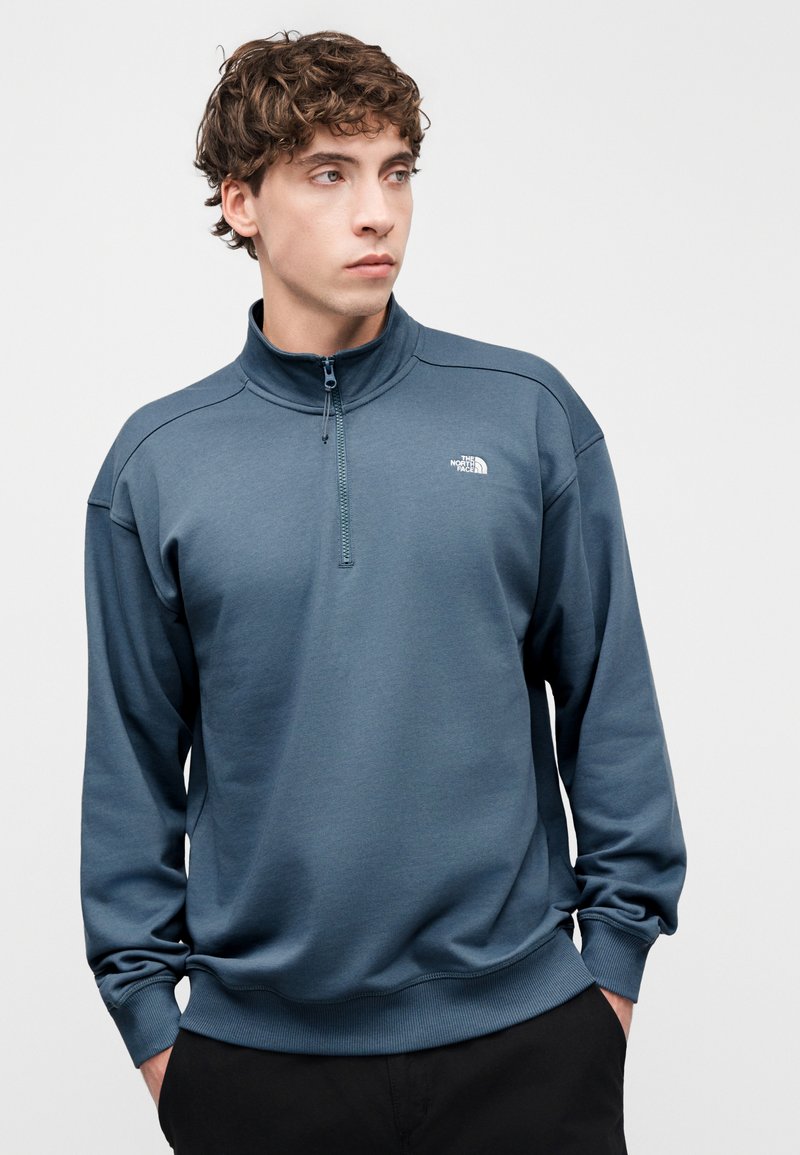 Blue quarter-zip fleece sweatshirt with long sleeves, ribbed cuffs and hem, featuring a small white logo on the left chest. Textured fabric.