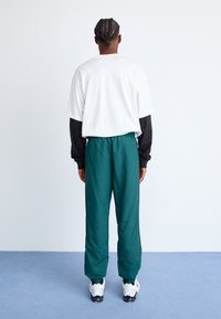 Hvit oversized t-skjorte lagdelt over en svart langermet skjorte, kombinert med grønne tapered bukser og hvite joggesko.