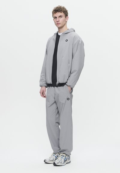 Coupe-vent gris clair avec fermeture éclair à l'avant, capuche et accent logo, associé à un pantalon gris assorti. Porté avec des chaussures de sport argentées.
