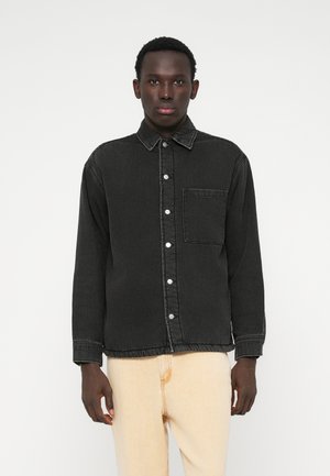 Camisa de denim preto com fecho de botões na frente, colarinho, um bolso no peito e bainha elástica. Apresenta costuras contrastantes e mangas compridas.